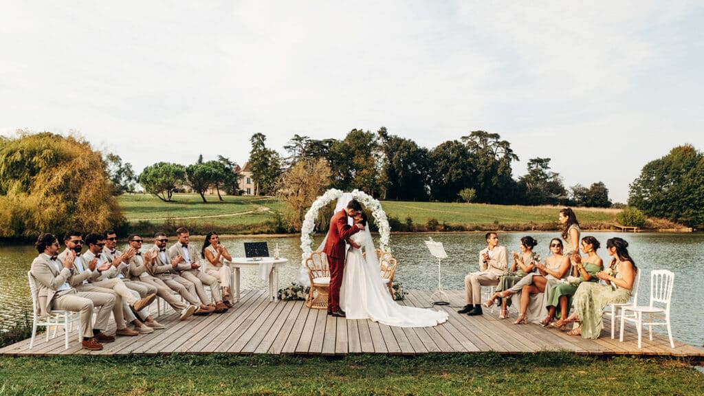 Mariage extérieur : Le baiser des mariés au bord du lac. Baiser des mariés sur un ponton de bois au bord d'un lac, sous une arche florale. Les invités en vert et beige applaudissent.